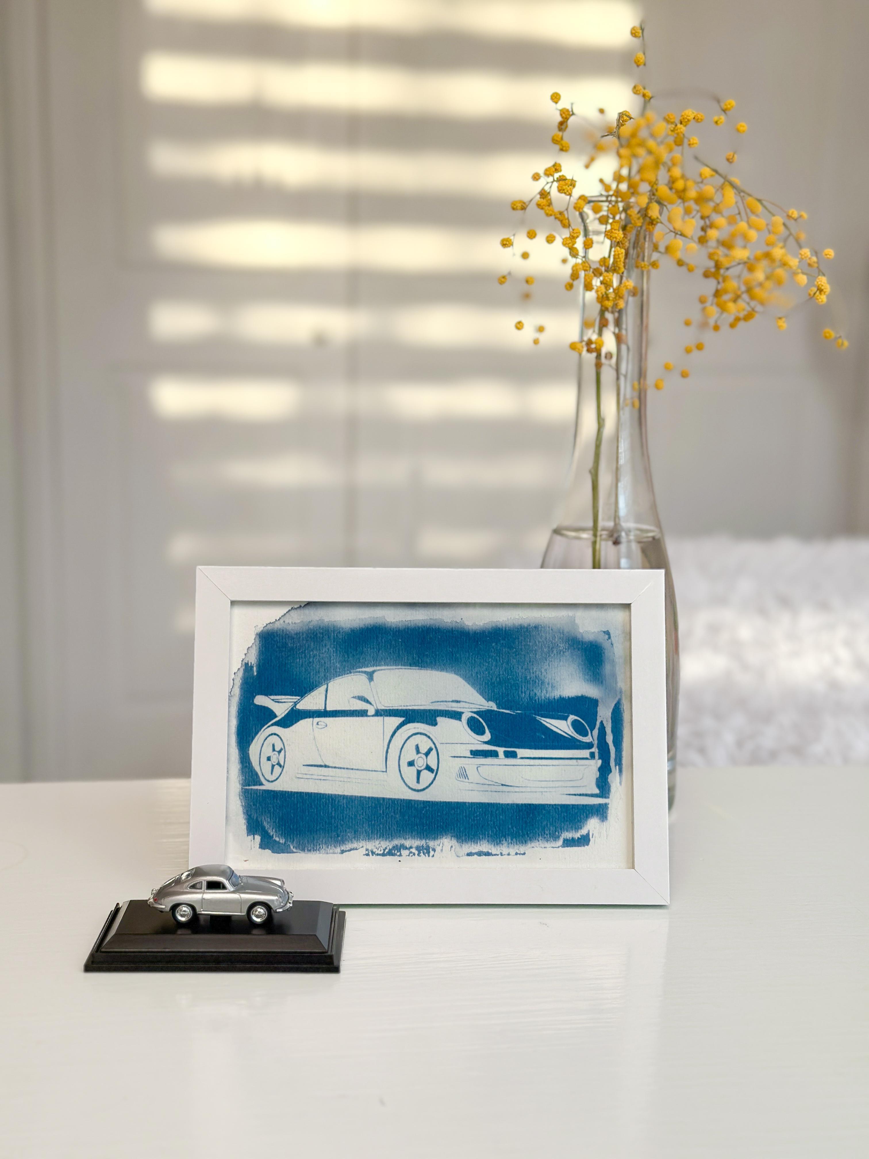 Cyanotype van een Porsche 911 in houten lijst, met miniatuurauto en vaas met gele bloemen op de achtergrond